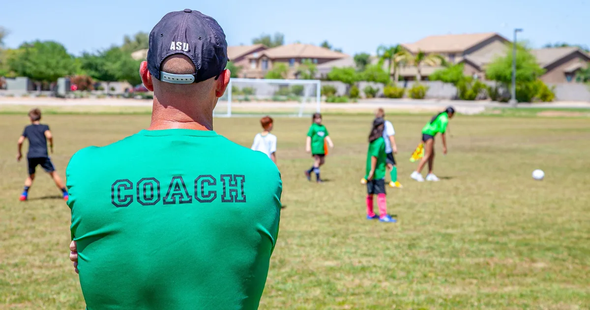 A youth sports coach standing on the field watching his soccer players at practice