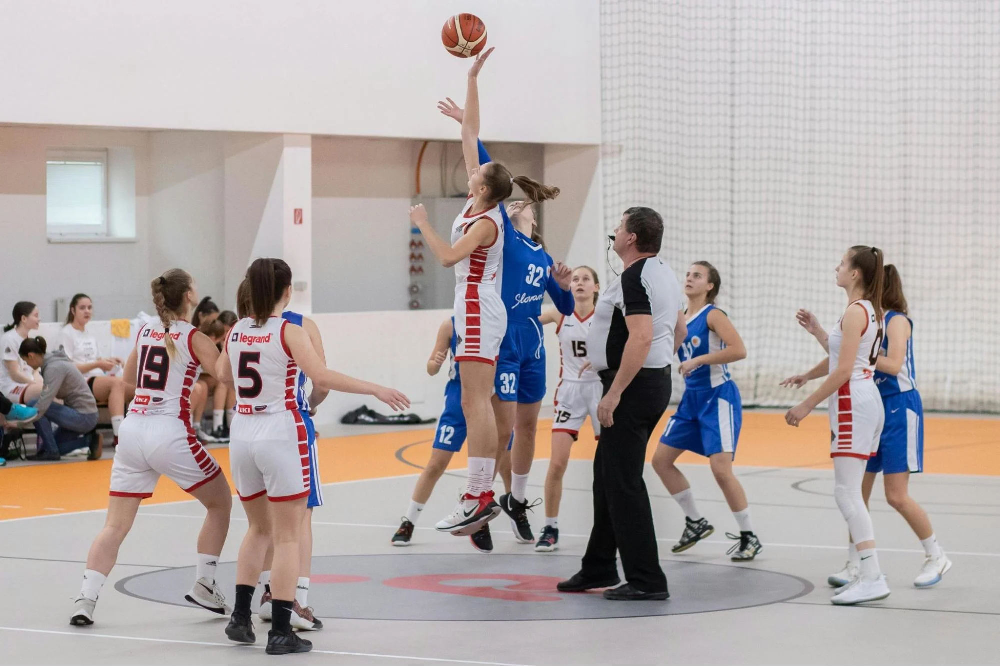 Girl’s basketball tip-off with two teams jumping for the ball at center court during a game.