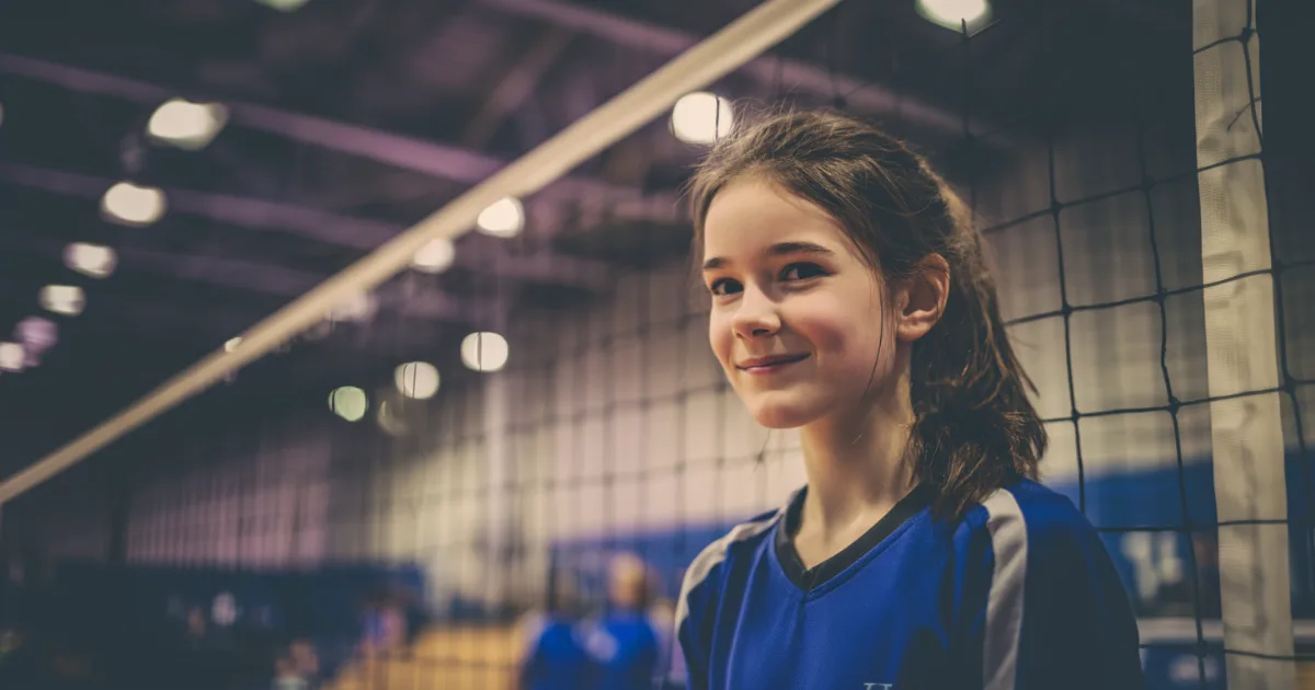 A youth volleyball player standing in front of the net smiling before a game