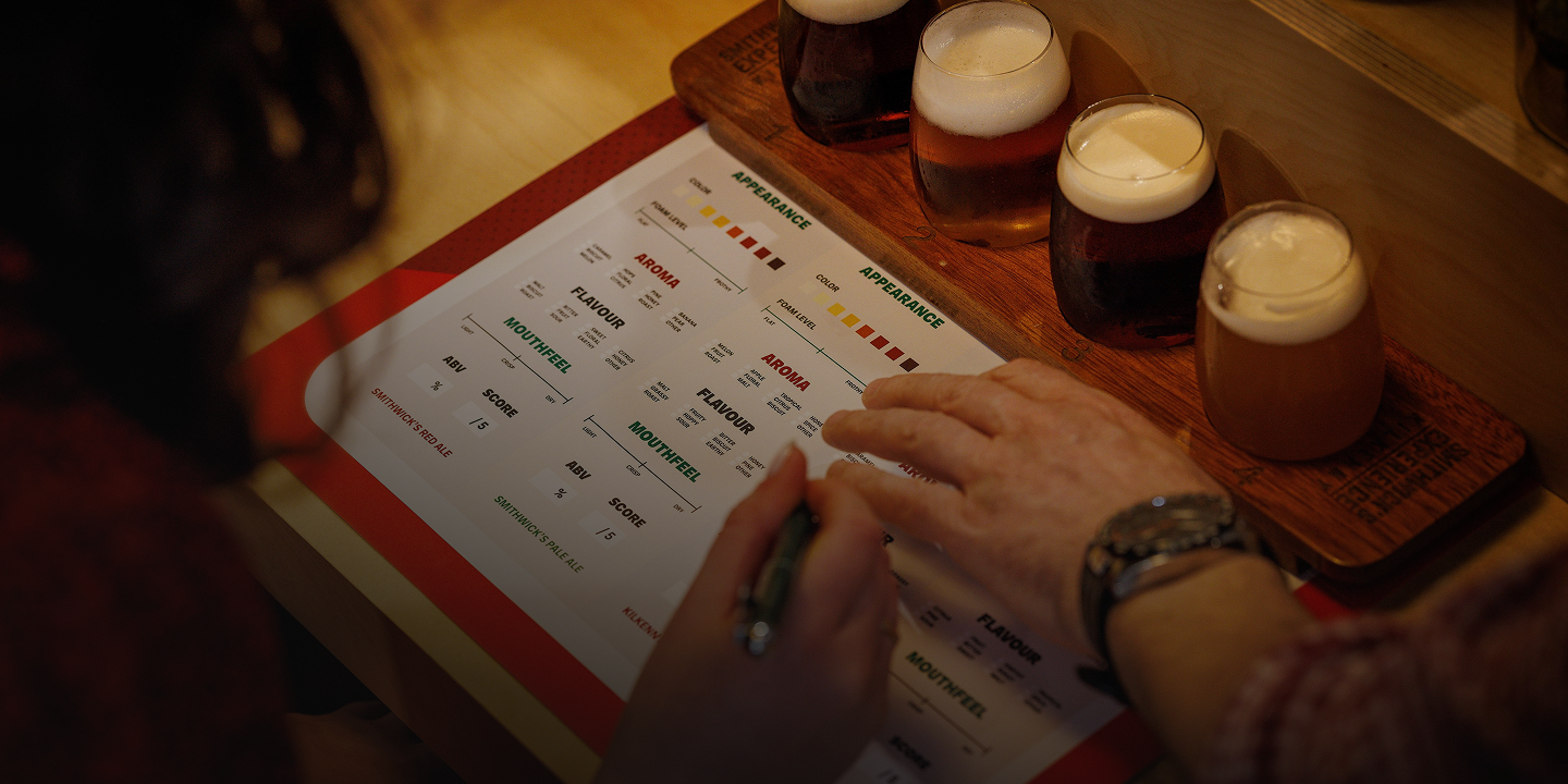 A hand fills out a Smithwick's beer tasting scorecard beside three glasses of ale on a wooden flight board.