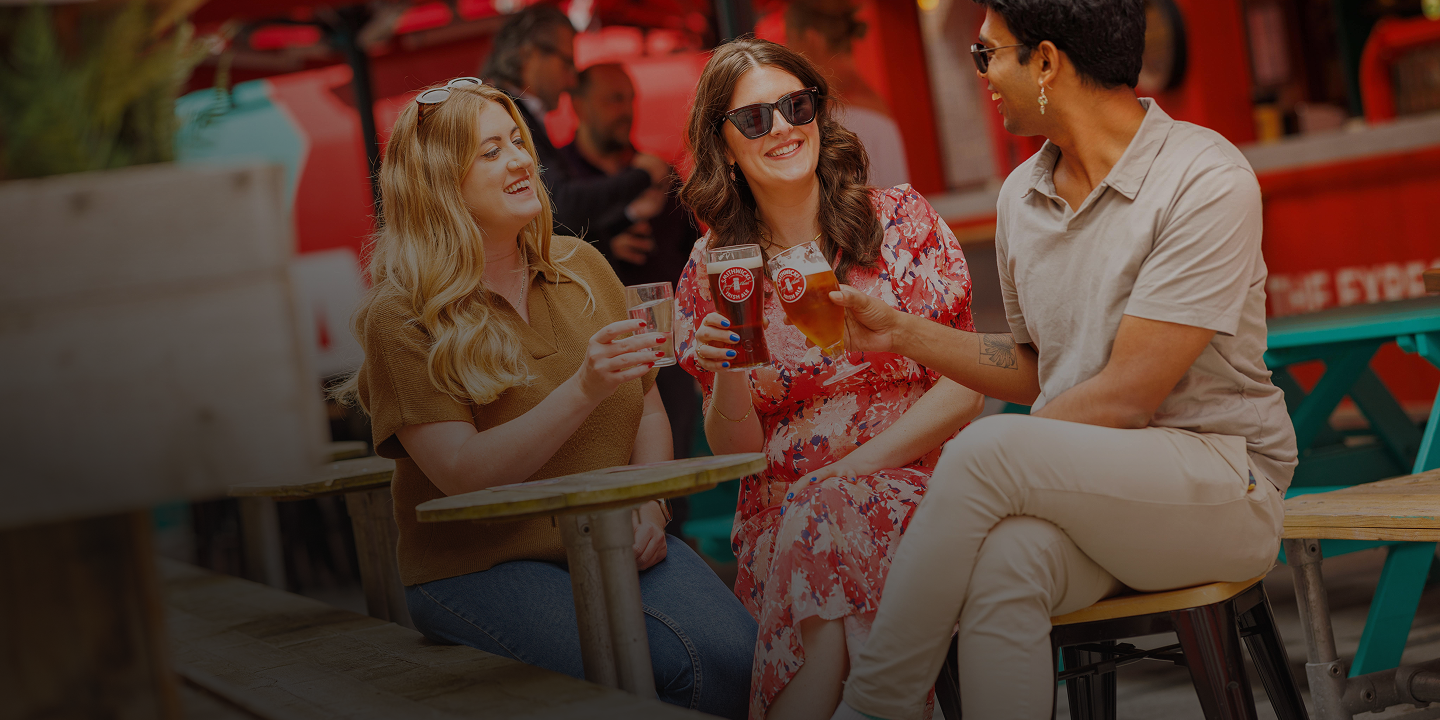 Three friends laugh and raise Smithwick's Irish Ale glasses at an outdoor table at the Smithwick's Experience.