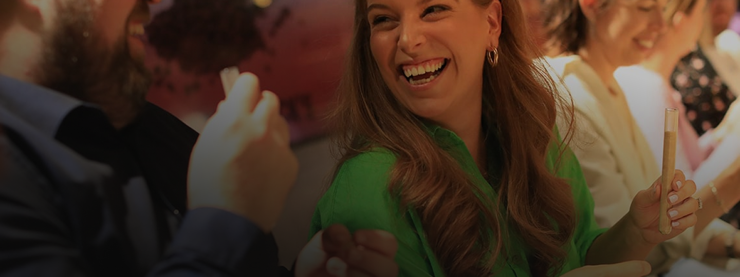 A laughing group of visitors enjoys an interactive tasting experience, with a woman in green holding a sample glass.