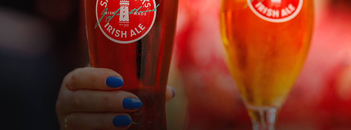 A hand with blue nail polish holding a pint glass of Smithwick's Irish Ale on a wooden table, with a second Smithwick's Irish Ale in a stemmed glass in the background.