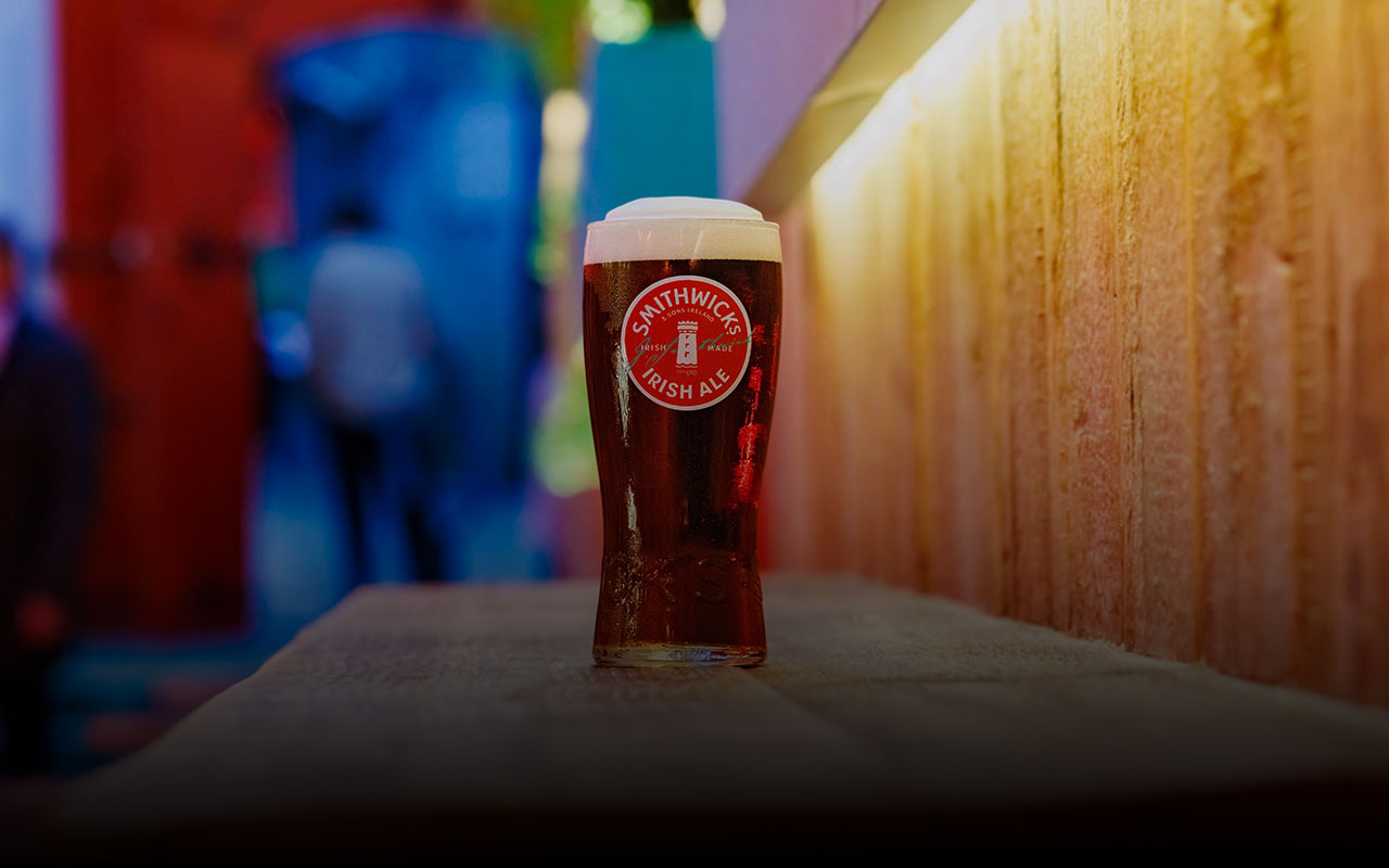 A hand with blue nail polish holding a pint glass of Smithwick's Irish Ale on a wooden table, with a second Smithwick's Irish Ale in a stemmed glass in the background.
