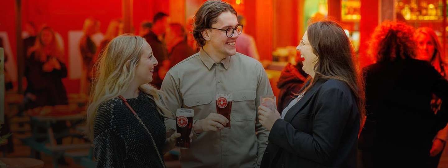 Three people holding drinks and talking in a lively bar with red lighting.