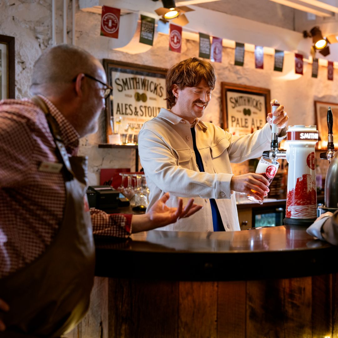 A smiling visitor pours a pint of Smithwick’s at a traditional pub bar while a staff member looks on and gestures. Warm lighting, branded beer taps, vintage Smithwick’s posters, and decorative bunting create a welcoming brewery-experience atmosphere.