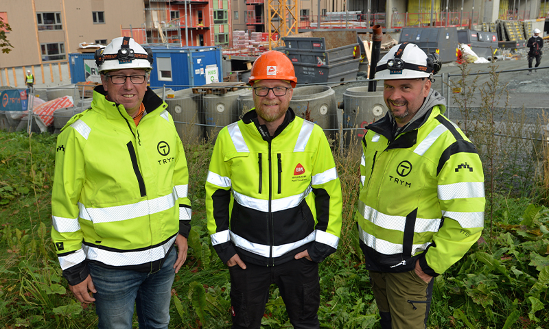 Tre byggearbeidere i neongule refleksjakker og hjelmer foran byggeplass med stillas og containere