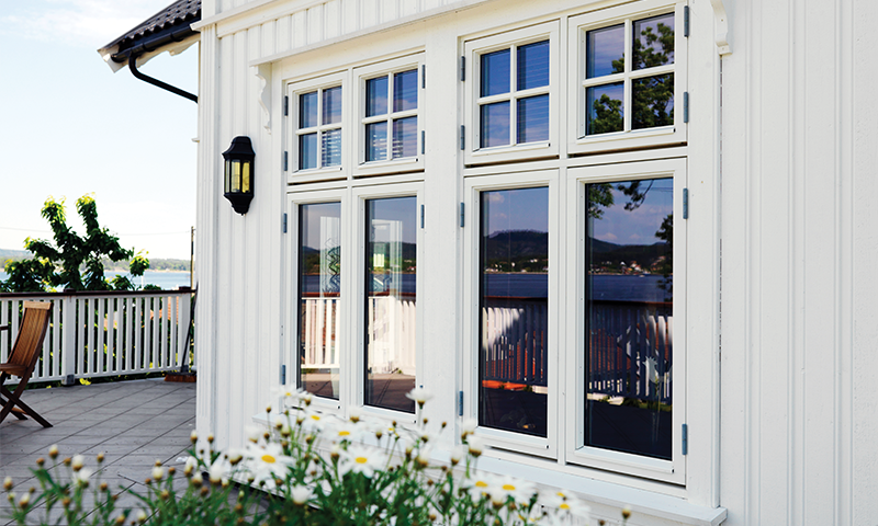 Hvitt trehus med store vinduer, blomstrende prestekrager og terrasse mot blå innsjø.