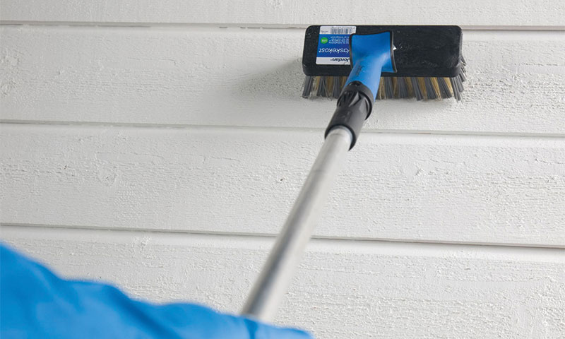 Hånd bruker blå skrubbebørste med teleskopskaft på hvit trepanelvegg.