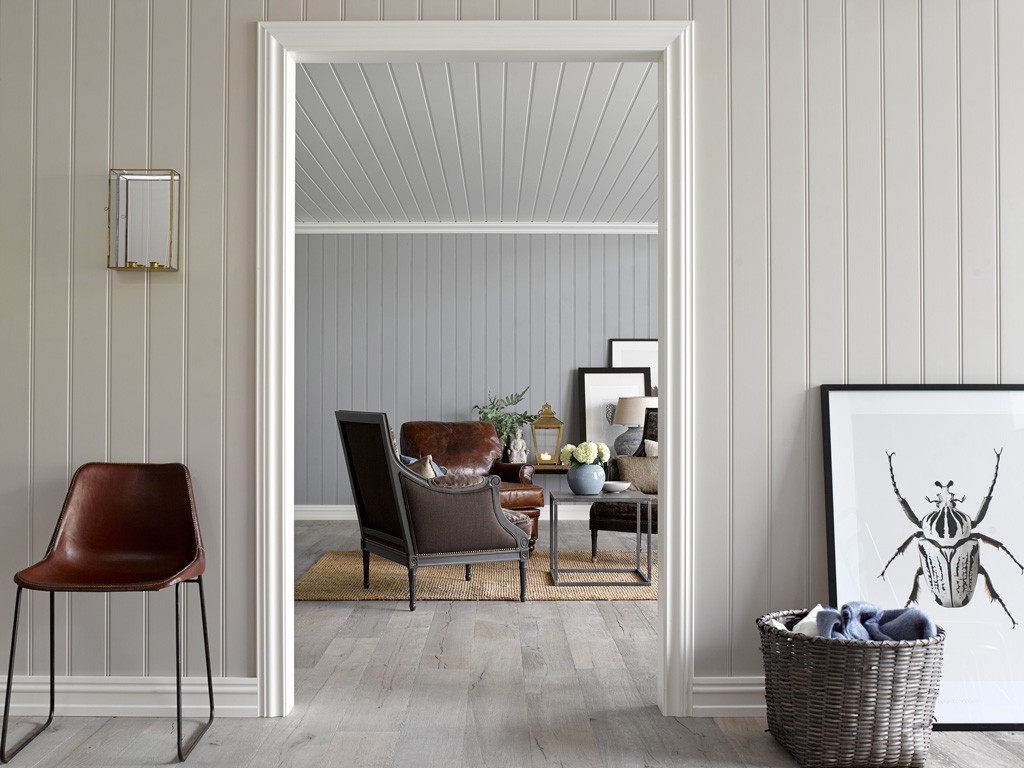 Stue gjennom døråpning: grå panelvegger, brun skinnstol, kaffebord, kurv med pledd, insektplakat.