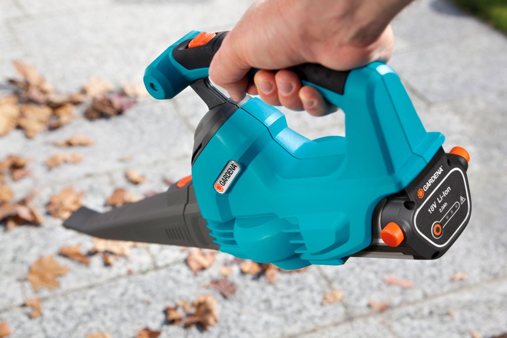 Hånd som holder turkis løvblåser med sorte og oransje detaljer over grå brostein med løv