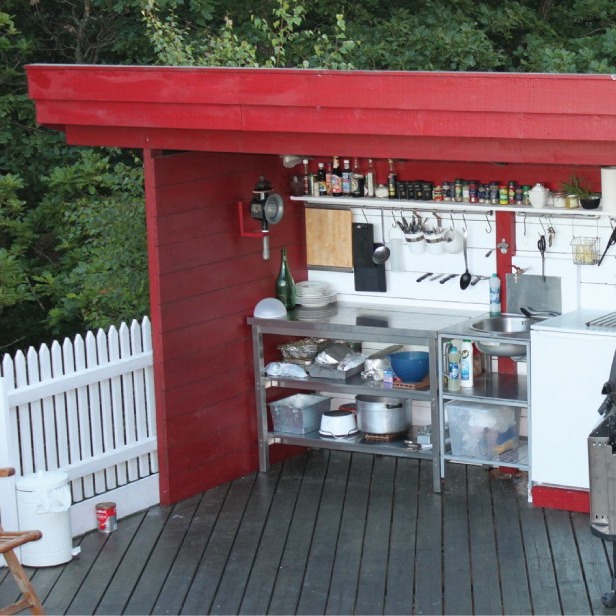 Utendørs rødt trekjøkken på mørk terrasse med stålbenk, gryter, krydder og hvitt gjerde.