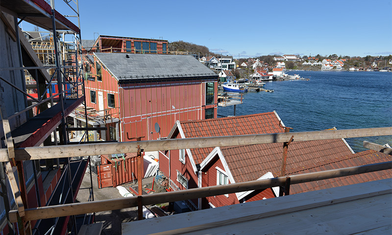 Stillaskant foran røde sjøboder under oppussing ved blått hav med båter og hvite hus.
