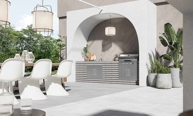 Utendørs spiseplass med hvite stoler, trebord, hengende lamper og grått grillkjøkken under bue