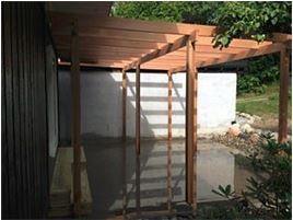 Trepergola foran hvit murvegg, skygger speiles i vannpøl, mørk tømmervegg til venstre