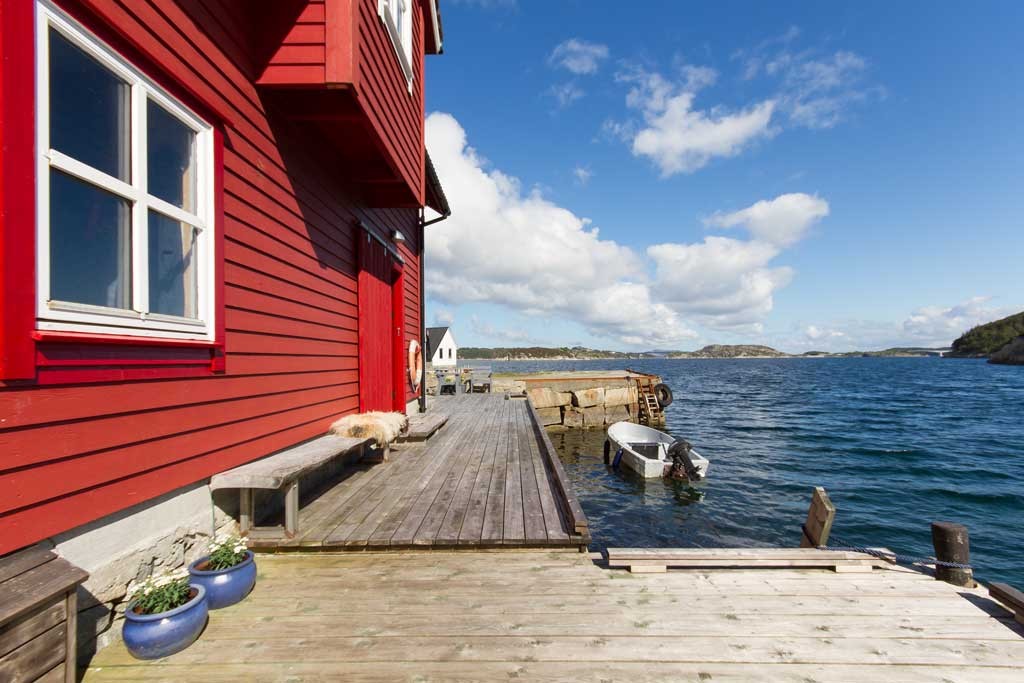 Rødt naust på trebrygge ved blått hav, lita båt og øyer under skyfri himmel