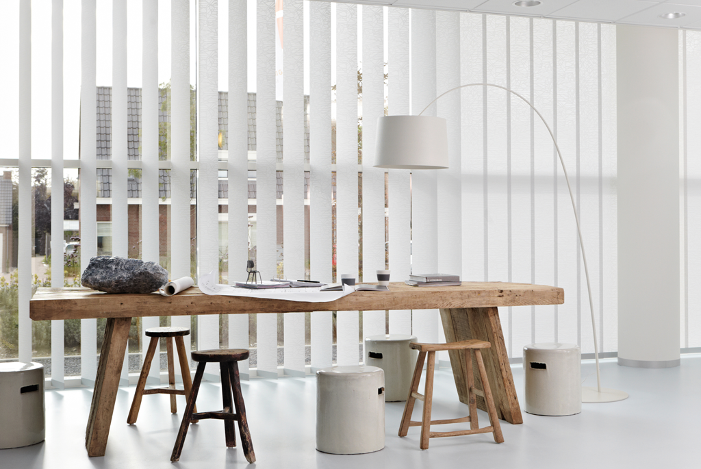 Lyst møterom med grovt trebord, hvite lameller, krakker, stein, tegninger og buegulvlampe