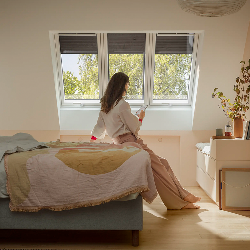 Person sitter på sengekanten i et lyst soverom med takvinduer, ser ut mot grønne trær i solskinn.