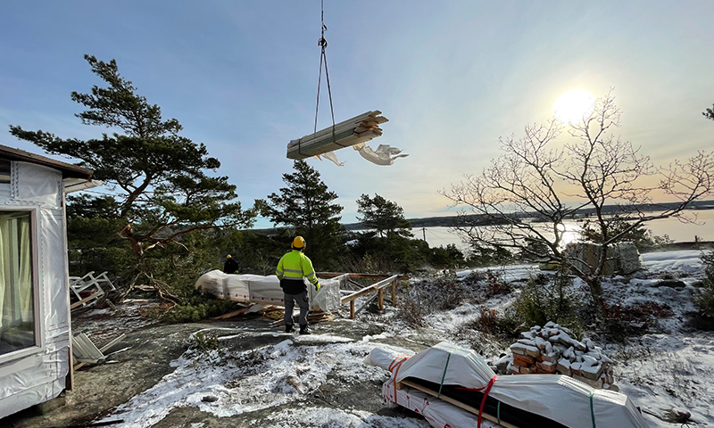 Byggearbeider i gul vest ser på kran som løfter trelast over snødekt fjellknaus ved kysten i sol