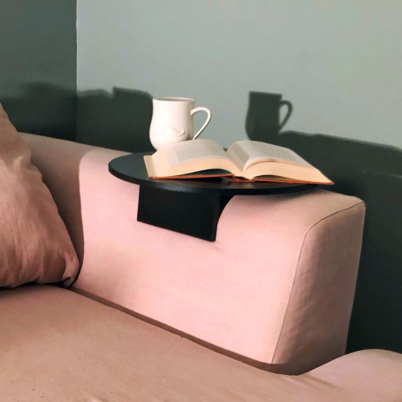 Åpen bok og hvit kopp på svart armtabell festet til rosa sofa foran grå vegg