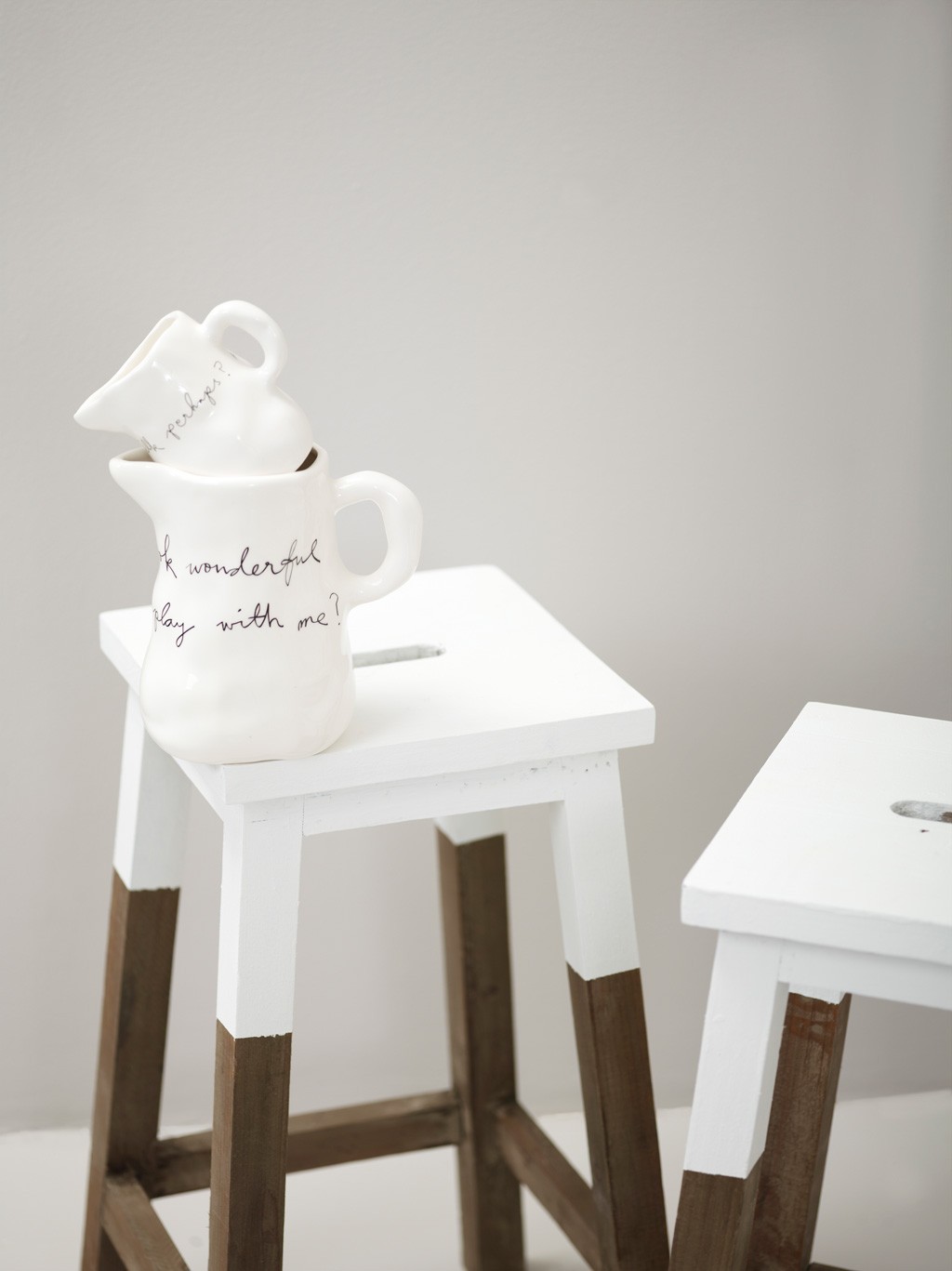 To hvite keramikkmugger med svart håndskrift stablet på hvit trekrakk med naturfargede bein.