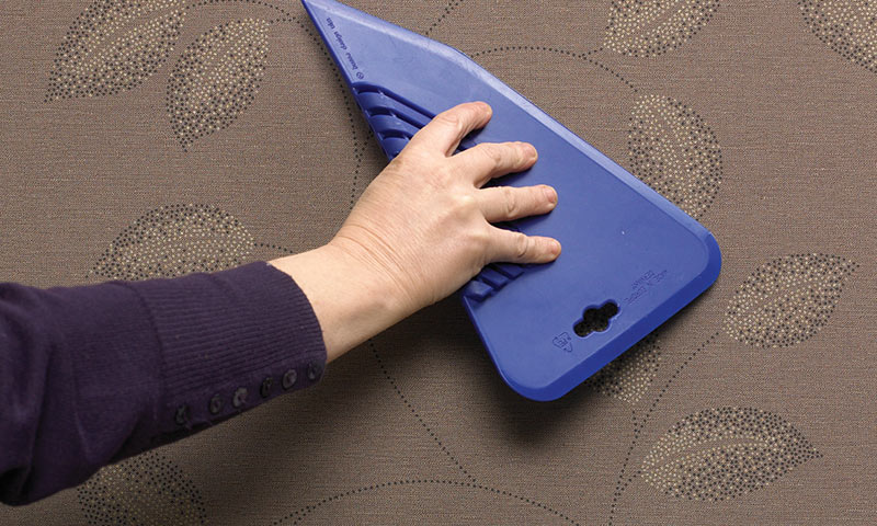 Hånd med blå plastskrape glatter brun tapet med prikkete bladmønster