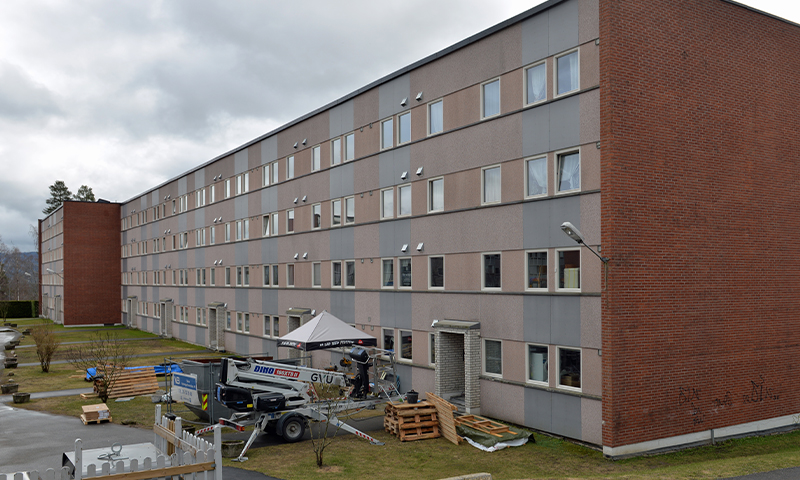 Fireetasjes boligblokk i beige-grå paneler med rød mur og byggeutstyr foran.