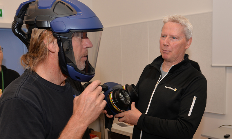 To menn innendørs, én får påmontert blått vernehjelm med visir og gassfilter.