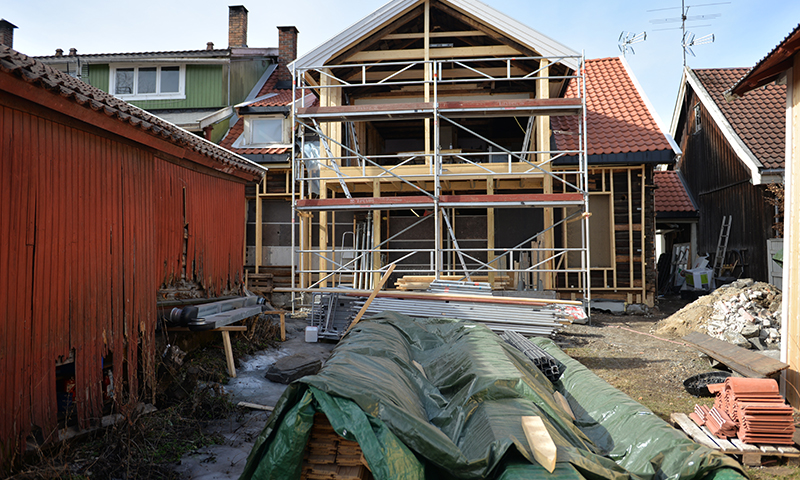 Halvferdig hus med stillas og materialer, grønn presenning og rød låve i bakgård.