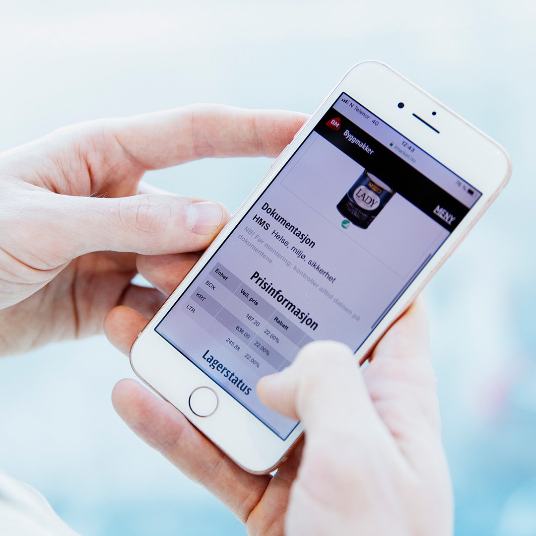 Nærbilde av to hender som holder hvit mobil med blå-hvit Byggmakker-nettside for pris- og HMS-info.