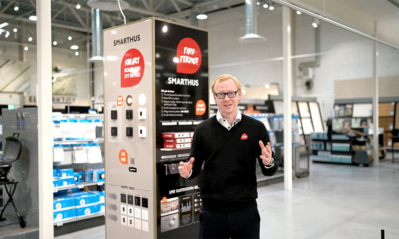 Ansatt i svart genser ved smarthus-stand med svart-hvit-rødt display i lys butikk