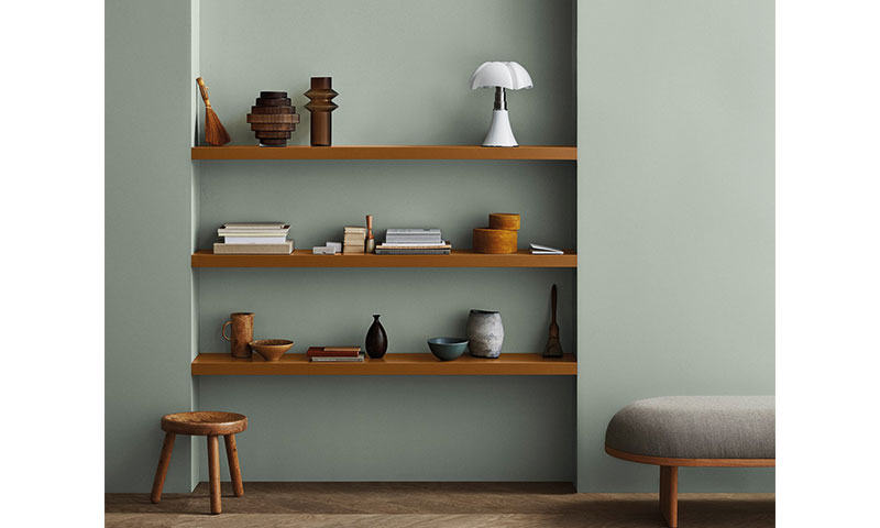 Grønn vegg med tre brune hyller med bøker, tre- og hvite dekorobjekter, skammel og grå benk