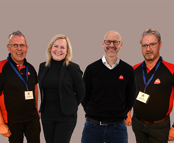 Fire ansatte foran grå bakgrunn i rød/svart bedriftsuniform, sort dress og genser