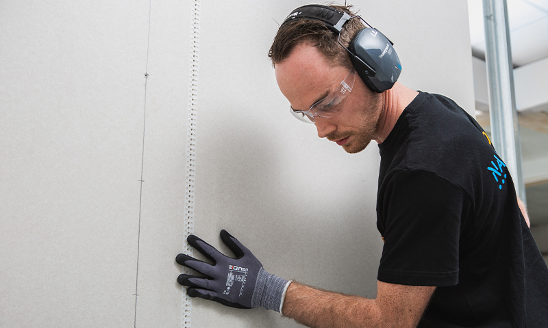 Bygningsarbeider med hørselsvern og hansker holder en lys grå gipsplate mot innvendig stenderverk.