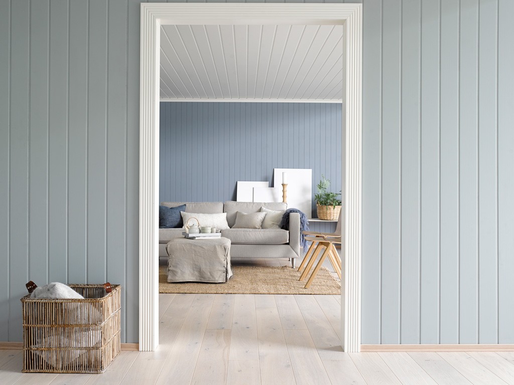Stue sett gjennom hvit dørkarm: blågrå panelvegger, grå sofa, jute­teppe, kurv med pledd