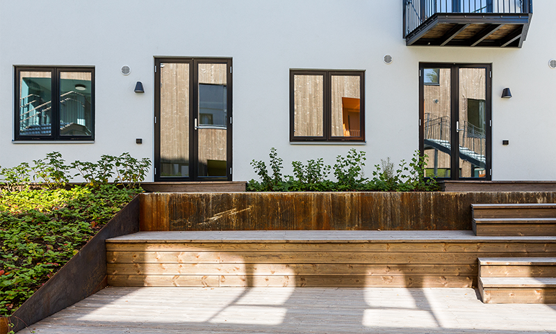 Hvit fasade med svartinnrammede glassdører og vinduer, tretrapp, balkong og grønn beplantning.