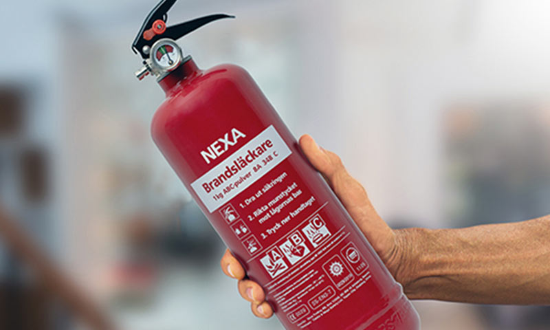 Hånd holder rødt Nexa ABC-pulver brannslukningsapparat med trykkmåler foran uskarp bakgrunn.