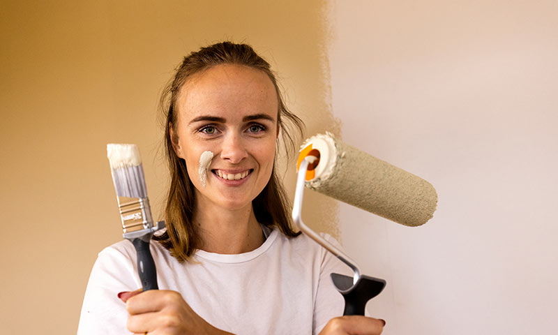 Person i hvit t-skjorte maler vegg med rull og pensel i beige tone.