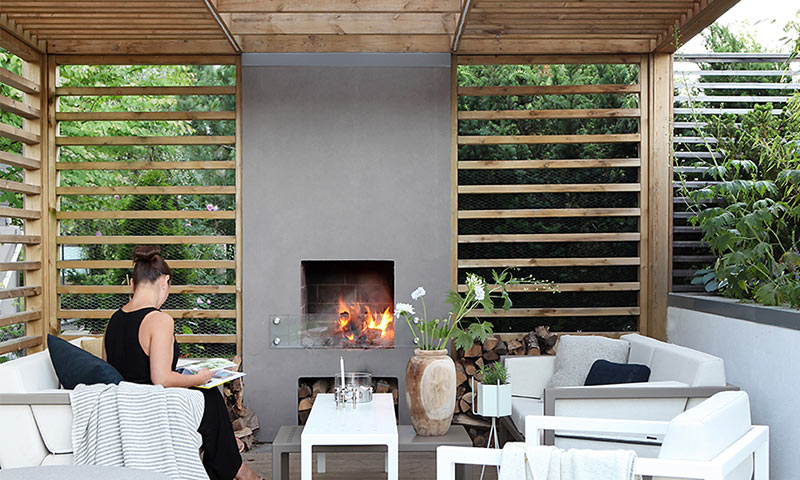 Utendørs lounge med trepergola, grå peis med ild, hvite sofaer, kvinne leser bok