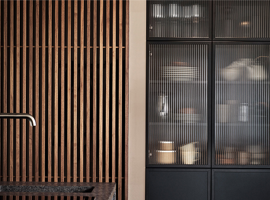 Moderne kjøkken med svart benkeplate, krom kran, trelameller og glasskabinett med servise.