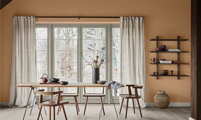 Spisebord med trestoler foran vindu, brun-oransje vegg, keramikk og vase med tørkede kvister.