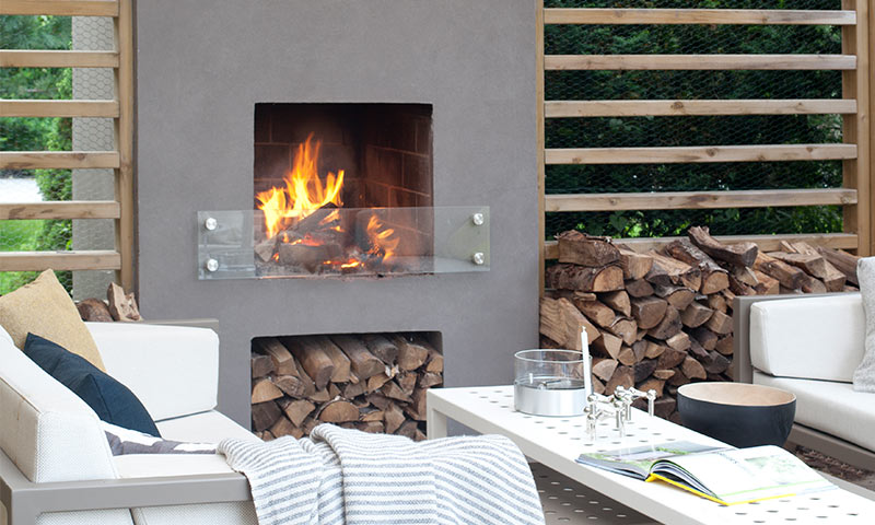 Utendørs sittegruppe med grå peis, flammer, vedstabel, hvite sofaer, trepanel og bord.