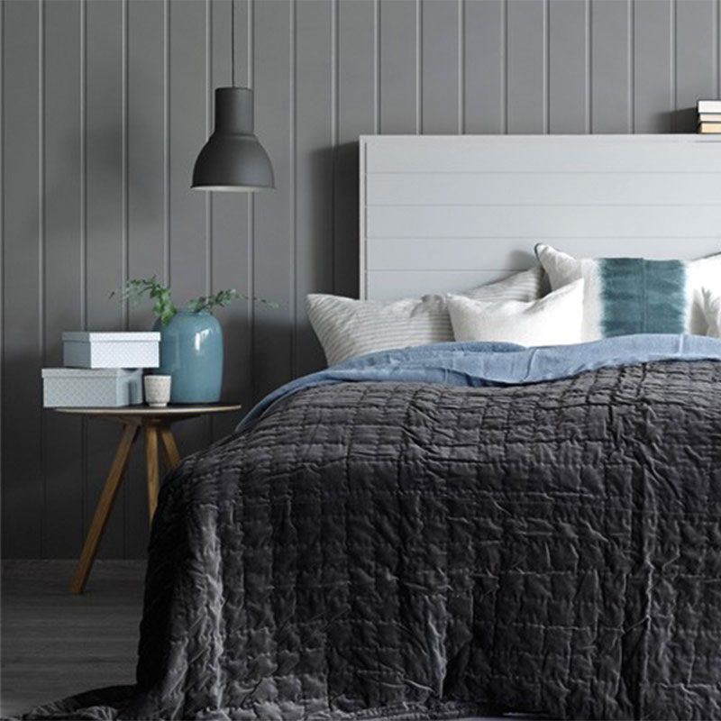 Soverom med dobbeltseng med mørkegrått teppe, blått pledd, hvite puter, sidebord med vase og lampe.