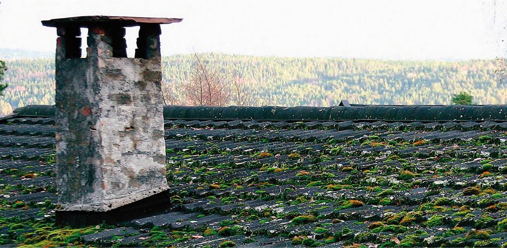 Grå steinpipe på svart tak med grønn mose, utsikt mot lys skog i bakgrunnen.
