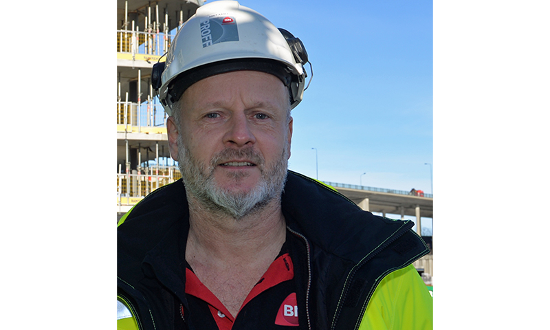 Mann i hvit hjelm og gul refleksjakke foran uferdig betongbygning under blå himmel.