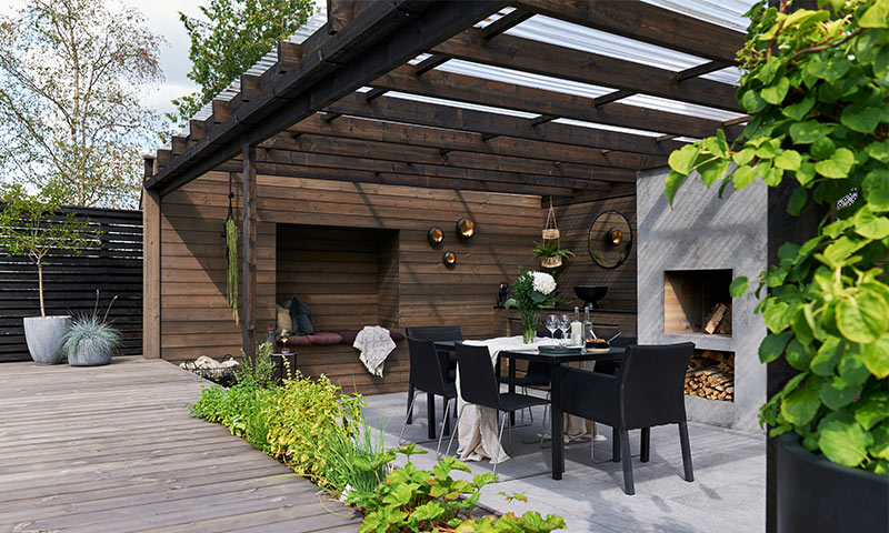 Uteplass med pergola i mørkt tre, svart spisebord med stoler, grå peis, grønne planter.