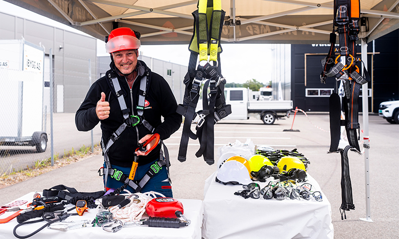 Person i svart jakke og rød hjelm gir tommel opp ved stand med gule hjelmer og seler under telt