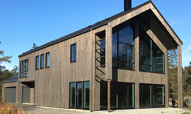Moderne to-etasjes trehus med mørk kledning, store vinduer, skråtak mot blå himmel