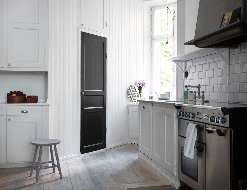 Lyst, hvitt kjøkken med svart dør, stålkomfyr, blomster ved vindu og lilla krakk.