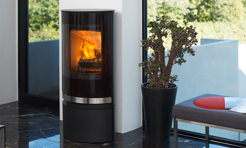 Svart sylinderpeis med flammer foran vindu, grønn plante i krukke og grå benk med rødt sete.